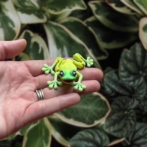 May include: A small, lime green frog figurine with a red mouth and black eyes. The frog is resting on a hand, with a blurred background of green and white leaves. The frog's limbs are outstretched, and it appears to be made of clay.