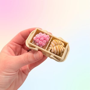 Puede incluir: Una peque&ntilde;a cesta beige dividida en dos compartimentos. Uno contiene un grupo de esferas rosas perladas y el otro dos objetos a rayas de color beige. La cesta se sostiene en una mano sobre un fondo pastel.