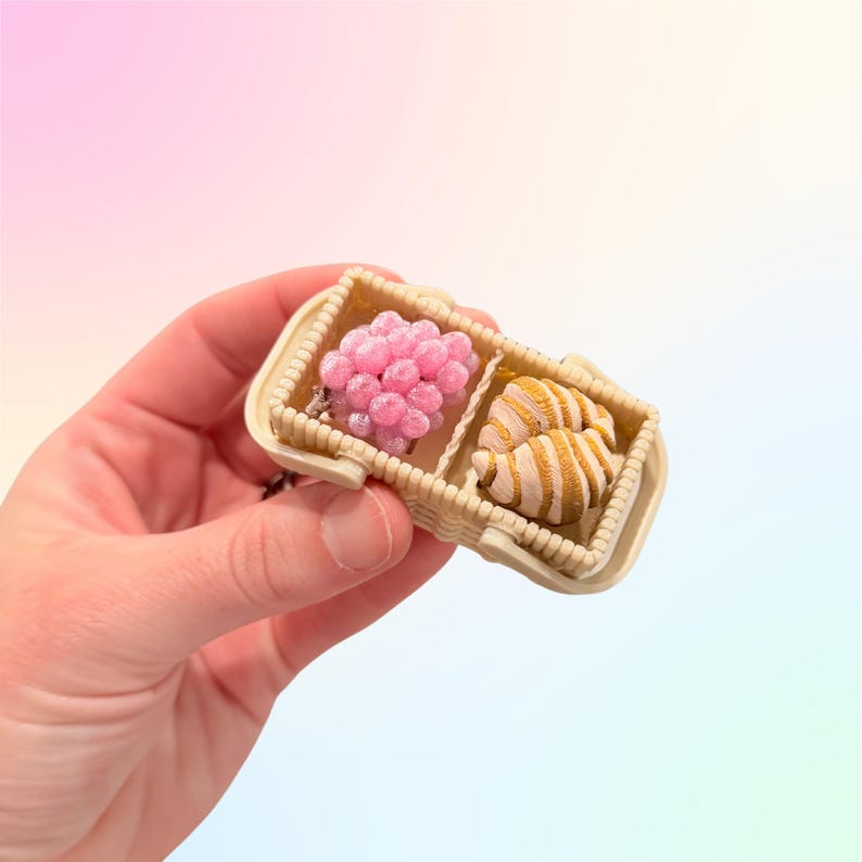 Puede incluir: Una peque&ntilde;a cesta beige dividida en dos compartimentos. Uno contiene un grupo de esferas rosas perladas y el otro dos objetos a rayas de color beige. La cesta se sostiene en una mano sobre un fondo pastel.