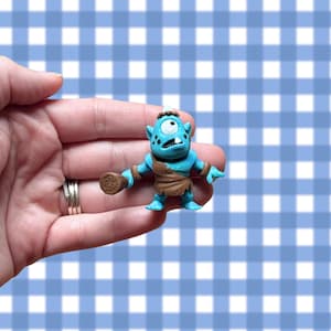 May include: A small, turquoise cyclops figurine with a single white eye, holding a brown club. The figurine has a brown loincloth and is set against a blue and white checkered background.