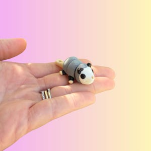 May include: A miniature opossum figurine with a gray body, black feet, and a white face. The figurine has a curled tail and is set against a gradient background of pink and yellow. The figurine is small enough to fit in the palm of a hand.