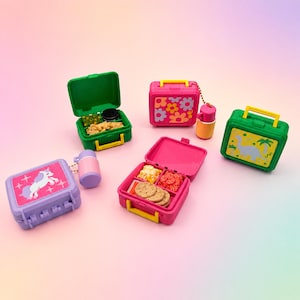 May include: A collection of five miniature plastic lunch boxes and matching thermoses in various colors including green, pink, and purple. Some boxes are open to reveal small food items like crackers and vegetables, while others are closed.
