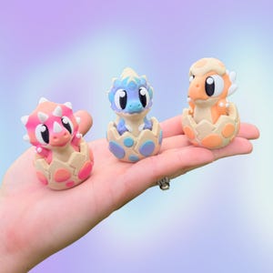 May include: Three small dinosaur figurines emerging from cracked eggs. The left dinosaur is pink with white spikes, the center one is blue with light blue accents, and the right one is orange with white spots. Each has large black eyes and a smooth finish.