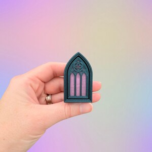 May include: A teal-colored, gothic-style window miniature with a pink interior, held in a hand. The window features an arched top and intricate detailing, including a quatrefoil design at the top. The miniature is likely a decorative item.