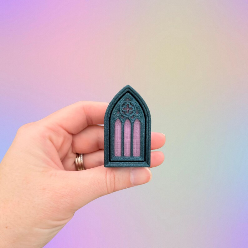 May include: A teal-colored, gothic-style window miniature with a pink interior, held in a hand. The window features an arched top and intricate detailing, including a quatrefoil design at the top. The miniature is likely a decorative item.
