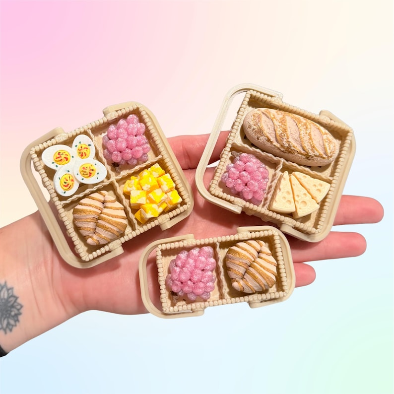 Puede incluir: Conjuntos de comida en miniatura en recipientes beige. Uno incluye huevos rellenos, esferas rosas, cubos amarillos y un cruas&aacute;n. Otro tiene pan, esferas rosas y cu&ntilde;as de queso. El tercer conjunto tiene esferas rosas y un cruas&aacute;n.