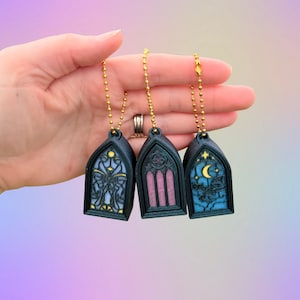 May include: Three gothic-style pendants with gold chains. Each pendant features a stained-glass design in shades of blue, pink, and yellow. One pendant has a butterfly, another has vertical lines, and the last has a moon and stars.