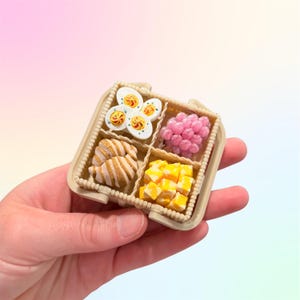 Puede incluir: Miniatura de fiambrera beige con cuatro compartimentos, cada uno lleno de peque&ntilde;os alimentos. Incluye huevos en rodajas, esferas rosas, cruasanes y cubos amarillos. La caja se sostiene en una mano sobre un fondo suave y colorido.