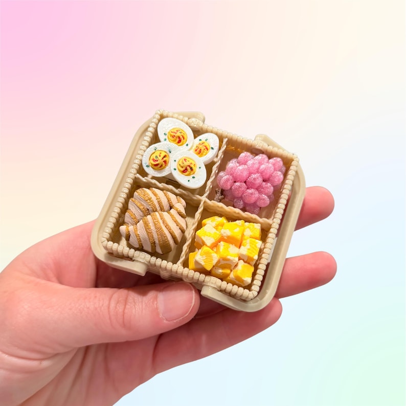 Puede incluir: Miniatura de fiambrera beige con cuatro compartimentos, cada uno lleno de peque&ntilde;os alimentos. Incluye huevos en rodajas, esferas rosas, cruasanes y cubos amarillos. La caja se sostiene en una mano sobre un fondo suave y colorido.