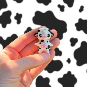 May include: A small, white cow figurine with black spots, a pink nose, and large, expressive eyes. The figurine is held in a hand against a backdrop of black and white cow print.