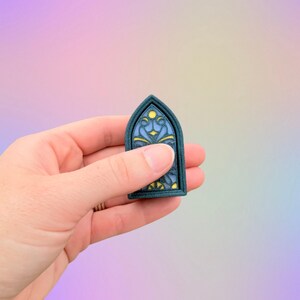May include: A small, teal-framed window-shaped charm with a stained-glass design. The interior features blue, yellow, and black details. The charm is held in a hand against a soft, pastel-colored background.
