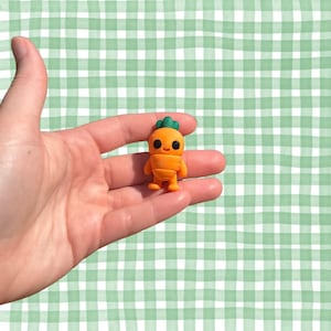 May include: A small, orange carrot-shaped figurine with a green leafy top. The figurine has black eyes and a smiling expression. It is held in a person's hand against a green and white checkered background.