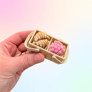 Puede incluir: Una cesta de picnic en miniatura con dos cruasanes y un racimo de uvas rosas. La cesta beige tiene un dise&ntilde;o tejido y un asa. Los cruasanes tienen rayas doradas.