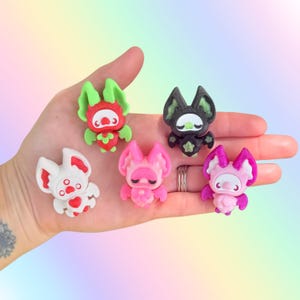 May include: Five small bat-shaped figurines in various colors including red with green ears, black with green accents, white with red details, solid pink, and magenta with light pink ears. Each bat features a distinct facial expression and design.