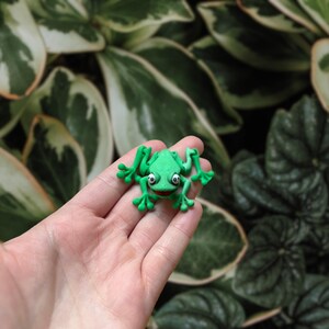 May include: A small, bright green frog figurine with white eyes and a red mouth. The frog is made of a smooth material and has its arms and legs outstretched. The figurine is held in a person's hand against a backdrop of green and white leaves.