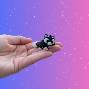 May include: A small, black and white cat figurine with large, expressive eyes and pink accents. The cat is lying down, with its tail and paws visible. The background is a gradient of purple and pink with white star-shaped sparkles.