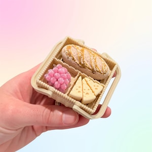 Puede incluir: Una peque&ntilde;a cesta de picnic con pan, uvas rosas y cu&ntilde;as de queso. La cesta beige tiene un asa y compartimentos. Los alimentos son detallados y realistas.
