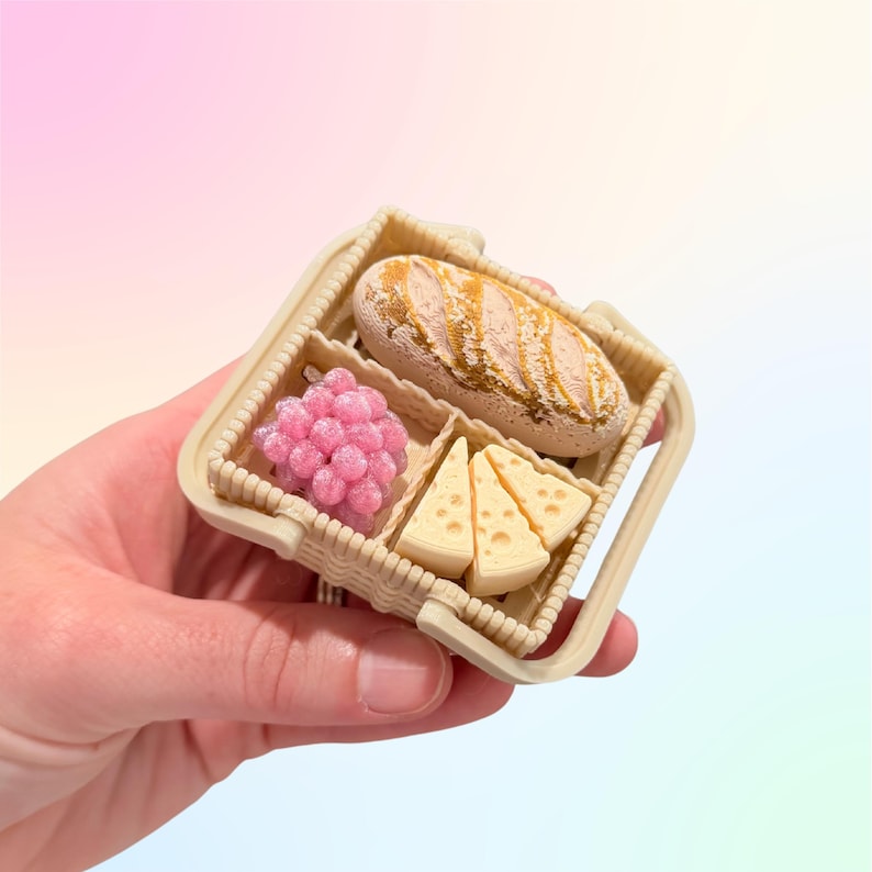 Puede incluir: Una peque&ntilde;a cesta de picnic con pan, uvas rosas y cu&ntilde;as de queso. La cesta beige tiene un asa y compartimentos. Los alimentos son detallados y realistas.
