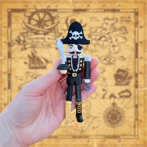May include: A pirate-themed nutcracker figurine. The figurine is dressed in a black coat with gold buttons, black pants, and a black hat with a skull and crossbones. It has a white mustache, an eye patch, and a sword. The hook is gray.