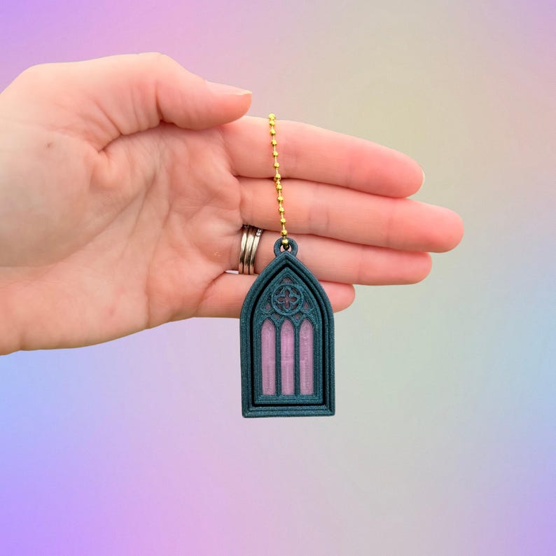 May include: A teal and pink gothic window-shaped ceiling fan pull with a gold chain. The pull has a detailed design with a floral emblem at the top and vertical lines inside the window frame.