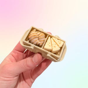 Puede incluir: Una cesta de picnic en miniatura llena de un cruas&aacute;n y cu&ntilde;as de queso. La cesta es de color beige con un dise&ntilde;o tejido. El cruas&aacute;n es de color marr&oacute;n claro con rayas marrones, y el queso es de color amarillo claro con peque&ntilde;os agujeros.
