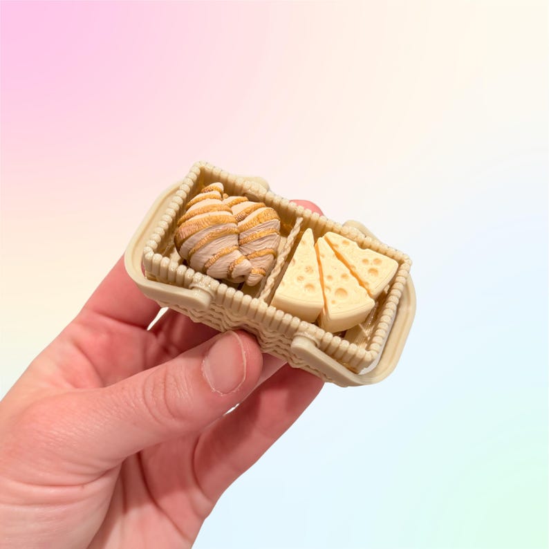 Puede incluir: Una cesta de picnic en miniatura llena de un cruas&aacute;n y cu&ntilde;as de queso. La cesta es de color beige con un dise&ntilde;o tejido. El cruas&aacute;n es de color marr&oacute;n claro con rayas marrones, y el queso es de color amarillo claro con peque&ntilde;os agujeros.