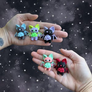 May include: A collection of handmade bat figurines in various colors, including blue, green, purple, pink, and red. Each bat has detailed wings and a unique design. The bats are displayed against a starry night background.