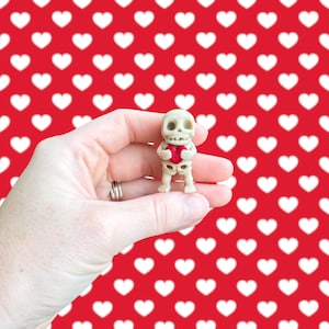 May include: A small, off-white skeleton figurine holding a red heart. The skeleton is detailed with visible ribs and skull features. The background is a repeating pattern of white hearts on a red surface.