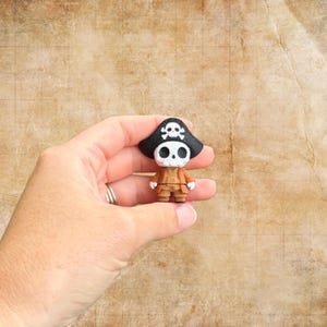 May include: A small, handcrafted pirate figurine. The figure has a skull-shaped head and wears a black hat with a skull and crossbones. It is dressed in an orange coat and brown pants. The figurine is held in a hand against a textured, tan background.