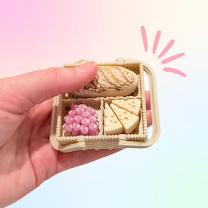 Puede incluir: Una cesta de picnic en miniatura beige con un pan, uvas rosas y cu&ntilde;as de queso. La cesta est&aacute; sostenida por una mano sobre un fondo pastel. El pan tiene una corteza dorada.