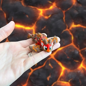 May include: A small, detailed figurine of a red and brown dragon with orange accents. The dragon is held in a hand against a background of lava. The figurine has large eyes and a friendly expression.