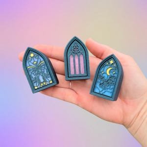 May include: Three teal-colored, arched window-shaped decorations with intricate designs. One features a butterfly, another has vertical pink bars, and the third showcases a moon and stars. The decorations are held in a hand.