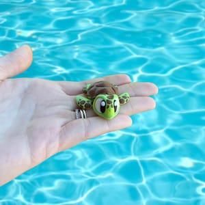 May include: A small, cartoon-style turtle figurine with a brown shell and green head and flippers. The turtle has large, expressive eyes and is held in a hand against a blue water background. The figurine is approximately 2 inches long.