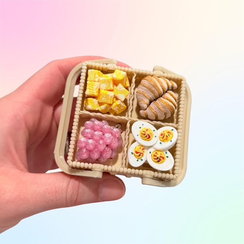 Puede incluir: Una cesta beige en miniatura dividida en cuatro secciones, cada una con un alimento diferente. Las secciones contienen comida amarilla en cubitos, un cruas&aacute;n, uvas rosas y huevos en rodajas. La cesta se sostiene en una mano.