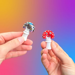 May include: Two mushroom-shaped bottle stoppers. One has a red cap with white spots, the other has a multicolored cap. Both have white stems. The stoppers are held against a gradient background of yellow, orange, purple, and blue.