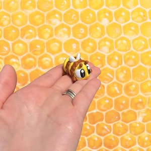 May include: A small, cartoonish bee figurine with a yellow body, brown stripes, and white wings. The bee has large, expressive eyes and is held in a hand against a honeycomb background. The figurine is approximately 1 inch in length.