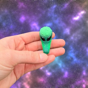 May include: A green alien figurine with large black eyes and a textured head. The alien is held in a hand against a blurred purple and blue galaxy background. The figurine is a vibrant green color.