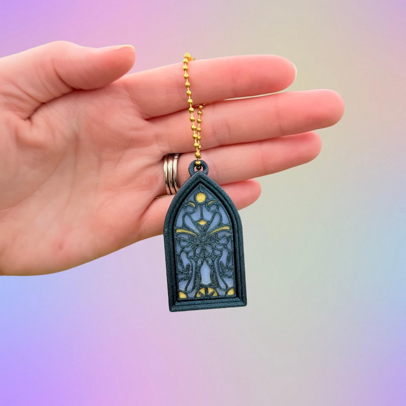 May include: A dark teal stained glass keychain with a gold chain. The keychain features a detailed design with blue and yellow accents. The design is reminiscent of a church window.