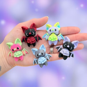 May include: Five small bat figurines in various colors including red and black, pink and green, light blue and grey, lavender and lime green, and black and lavender. Each bat has large eyes, pointed ears, and small wings, crafted with a textured finish.