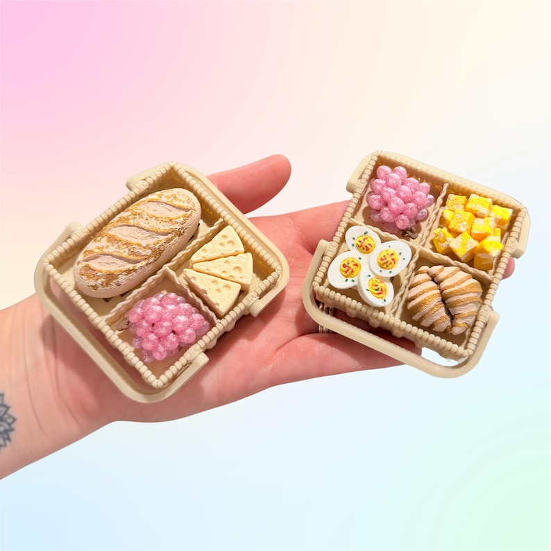 Puede incluir: Miniaturas de cestas de picnic llenas de alimentos. Una cesta contiene una barra de pan, cu&ntilde;as de queso y uvas rosas. La otra cesta tiene uvas, alimentos amarillos en dados, huevos decorados y cruasanes. Las cestas son beige.