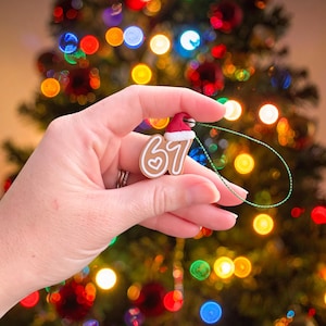 May include: A gingerbread-style Christmas ornament in the shape of the number 67, with a heart detail and a Santa hat. The ornament is held in front of a blurred Christmas tree with colorful lights.