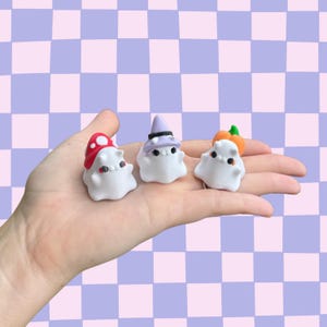 May include: Three small, white ghost figurines with black dot eyes and various hats. One ghost wears a red mushroom cap, another a purple witch hat, and the third a pumpkin. The background is a checkered pattern of lavender and white.