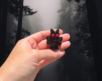 May include: A small, black figurine of a creature with red eyes and small wings. The figurine is held in a hand against a backdrop of a misty forest. The figure has a stylized design.