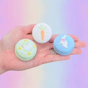 May include: Three pastel-colored macarons are held in a hand. One is light green with a white chick design, another is white with a carrot, and the last is light blue with a bunny. The macarons have a spiral design.