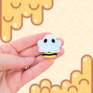 May include: A small, white and yellow bee-themed figurine with black eyes and pink cheeks. The figurine has a white top with small wings and a yellow and black striped bottom. The background features a honey-themed pattern.