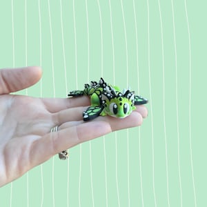May include: A small, green and black dragon figurine with white accents. The dragon has large, expressive eyes and is lying on its belly. The wings are a butterfly design. The figurine is held in a hand against a light green background.