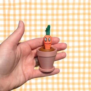 May include: A whimsical orange carrot with a smiling face and green top sprouts from a brown base inside a small, pink flower pot. The pot has a matching saucer. This decorative item is a fun, playful addition to any home or garden.