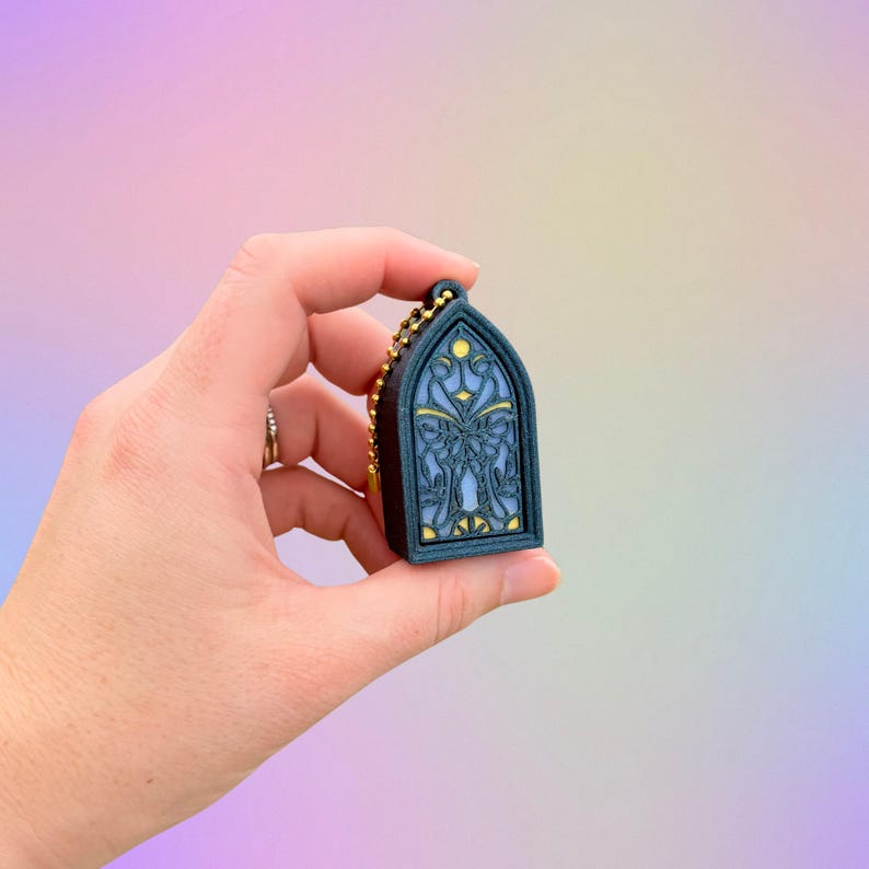May include: A dark teal-colored keychain shaped like a stained-glass window. The window design features a butterfly and floral pattern in shades of blue and yellow. The keychain has a gold-colored chain.