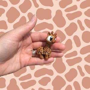 May include: A small, handcrafted giraffe figurine with a brown and tan spotted pattern. The giraffe has large, expressive eyes and small, rounded ears. The figurine is held in a hand against a background with a giraffe-spot pattern.