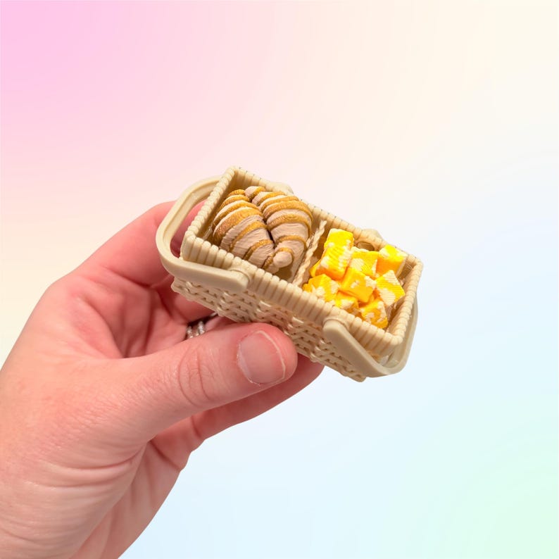 Puede incluir: Una peque&ntilde;a cesta de picnic beige con dos cruasanes y una porci&oacute;n de queso en cubos. Los cruasanes son dorados y hojaldrados, y el queso es amarillo. La cesta tiene un asa.